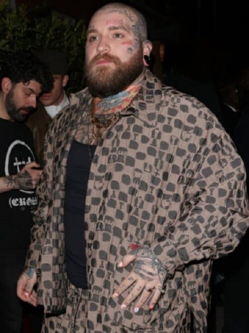 A bearded man with tattoos on his hands and face wears a Teddy Swims Brown Suit, a patterned brown oversized outfit paired with a black shirt. He accessorizes with layered necklaces and a colorful scarf. Captured at a nighttime event, he exudes a confident and stylish cyberpunk-inspired look.