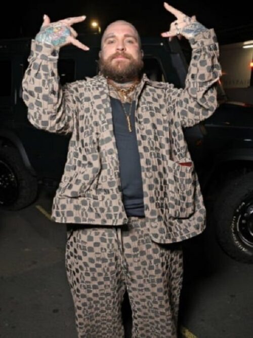 A bearded man with tattoos on his hands poses outdoors at night, wearing a Teddy Swims Brown Suit, a matching brown patterned outfit with a black shirt underneath. He holds up both hands in a rock-inspired gesture, with a confident expression. A dark-colored vehicle is visible in the background.