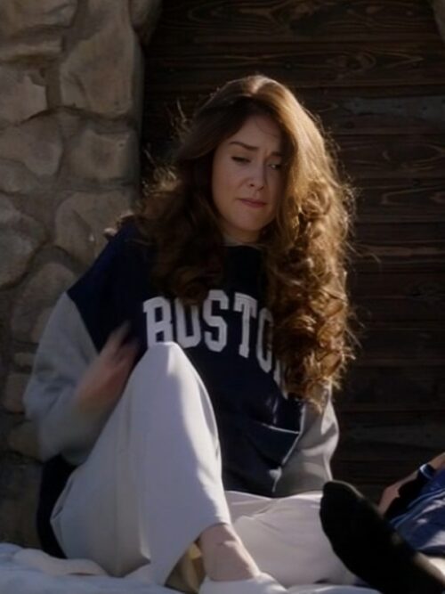 A woman with voluminous, wavy brown hair and a slightly uneasy expression is seated outdoors on a stone surface. She is wearing a The Royal We 2025 Mallory Jansen Sweatshirt with "BOSTON" in bold white letters on the front, paired with loose white pants and white slip-on shoes. The relaxed, casual look reflects a cozy and sporty aesthetic.