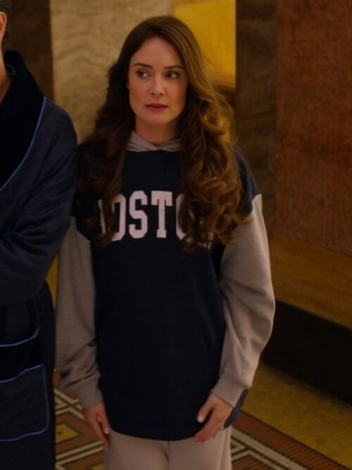 A woman with long, wavy brown hair and a concerned expression is wearing a The Royal We 2025 Mallory Jansen Sweatshirt with "BOSTON" printed across the chest. The oversized hoodie features a hood and full-length sleeves with contrasting gray fabric. The setting appears to be indoors with warm lighting and a partial view of another person in a robe beside her.