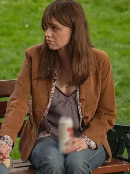 A woman with shoulder-length brown hair sits on a park bench wearing a Toxic Town 2025 Aimee Lou Wood Brown Blazer over a dark-colored camisole with lace trim. She has a serious expression as she gazes to the side. The blazer features button details and a floral-patterned inner lining visible at the cuffs and lapel. She is also wearing blue jeans and holding a blurred beverage can in her hand. The background is grassy and slightly blurred.